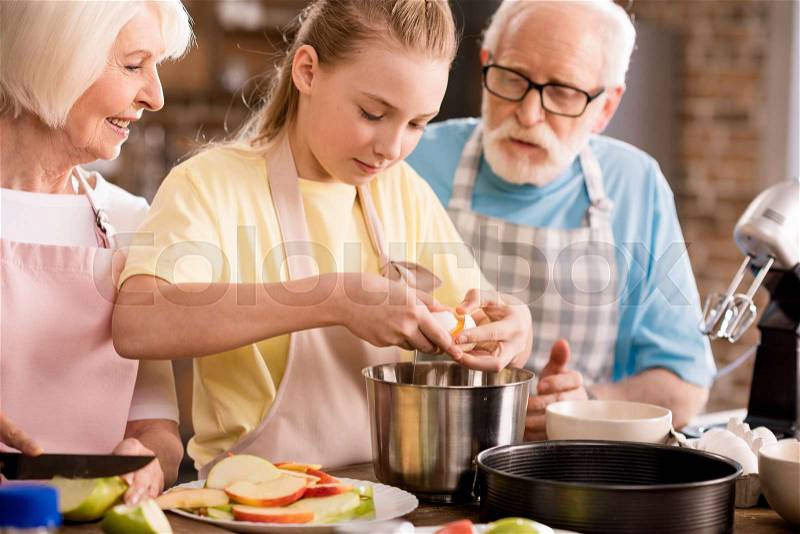 Happy grandparents with grandchild cutting apples and preparing dough for apple pie on kitchen table, stock photo