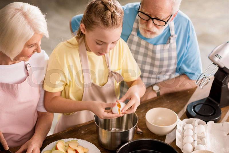High angle view of grandparents with granddaughter cutting apples and preparing dough for apple pie on kitchen table, stock photo
