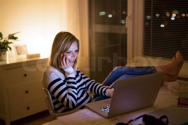 Young beautiful woman sitting at desk at night, holding smartphone, making phone call, working on laptop, stock photo