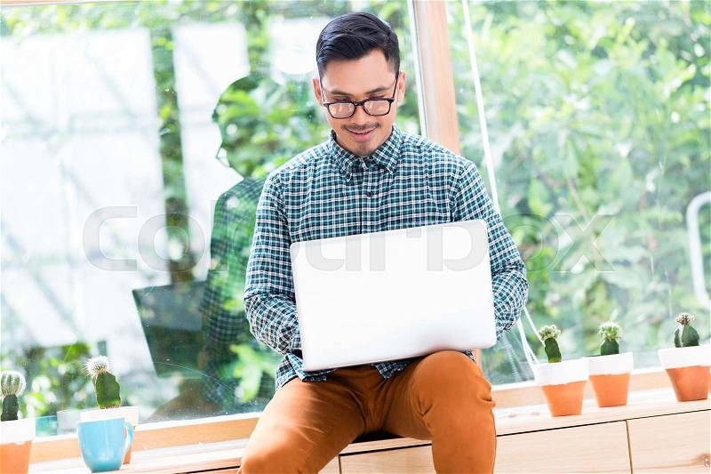 Relaxed young Asian employee working on laptop while sitting on a wooden drawer in the office, stock photo