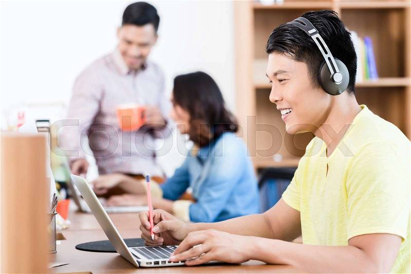 Side view portrait of a happy young Asian employee using headphones while watching a video presentation on laptop in the office, stock photo