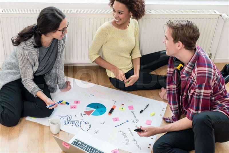 Three young people analyzing pie chart and writing observations on a large paper sheet during brainstorming session, stock photo
