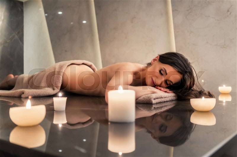 Scented round candle next to young woman on marble massage table during aromatherapy at luxury spa and wellness center, stock photo