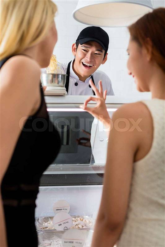 Young shop assistant laughing at a woman customer as she makes a perfect gesture with her hand as he weighs grated cheese, stock photo
