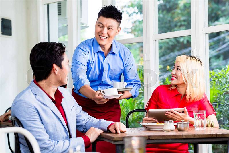 Happy Asian waiter serving a stylish couple coffee as they sit at a table in a restaurant enjoying a meal, stock photo