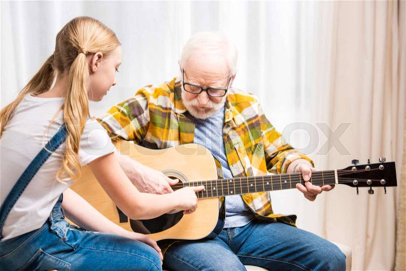 Senior man in eyeglasses teaching cute little granddaughter playing acoustic guitar, stock photo