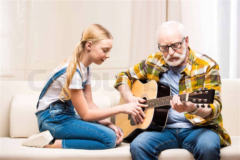 Senior man in eyeglasses teaching cute little granddaughter playing acoustic guitar, stock photo