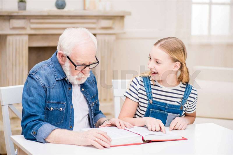 Smiling girl and grandfather in eyeglasses reading book together at home , stock photo