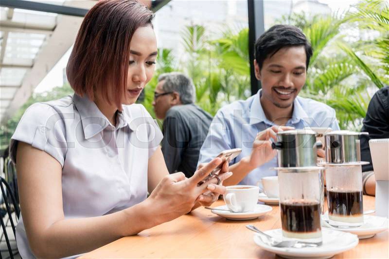 Three young Asian friends smiling while using electronic devices connected to the wireless internet network of a modern coffee shop, stock photo