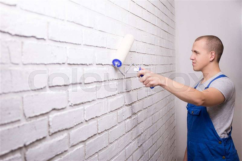 Young man painter in uniform painting white brick wall with paint roller, stock photo