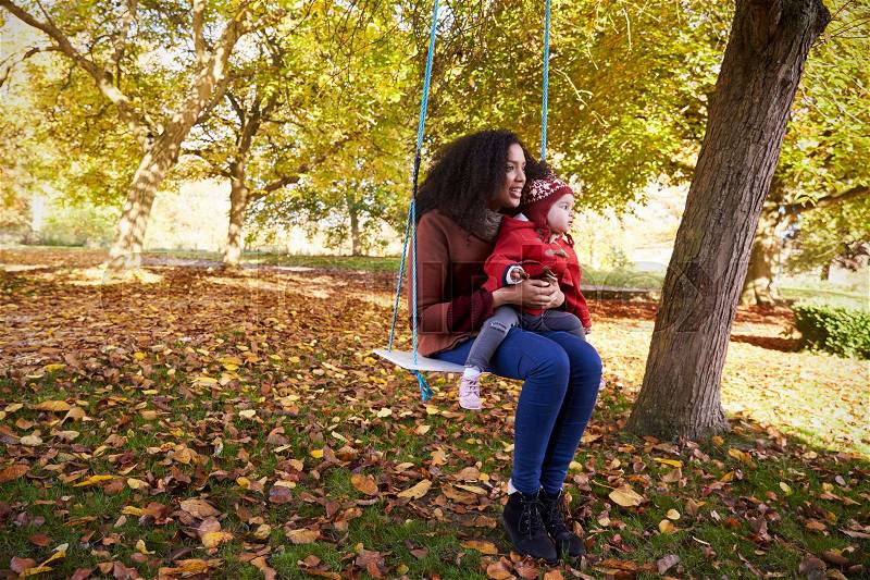 Mother With Daughter Playing On Tree Swing In Autumn Garden, stock photo