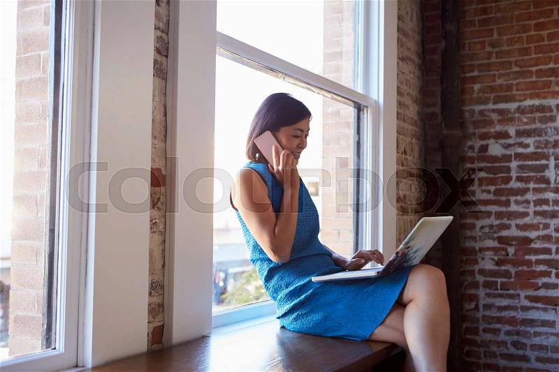 Businesswoman By Window Using Mobile Phone And Laptop, stock photo