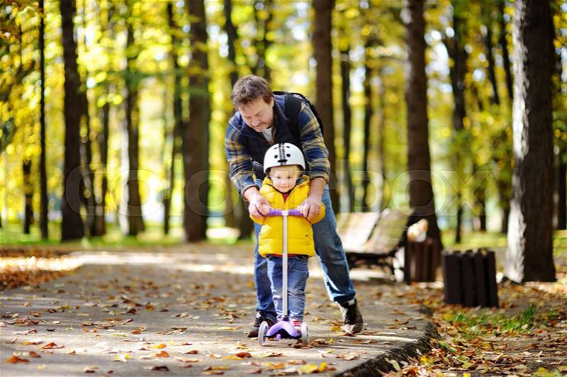 Middle age father showing his toddler son how to ride a scooter in a autumn park. . Active family leisure. Child in helmet. Safety, sports, leisure with kids concept, stock photo