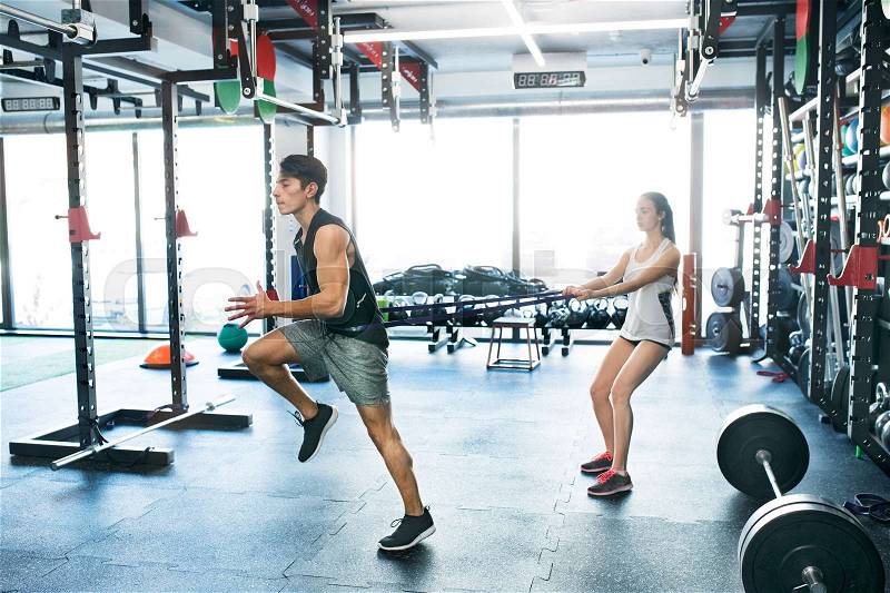 Strong man using a resistance band in his exercise routine. Young fit couple performs fitness exercise in modern crossfit gym, stock photo