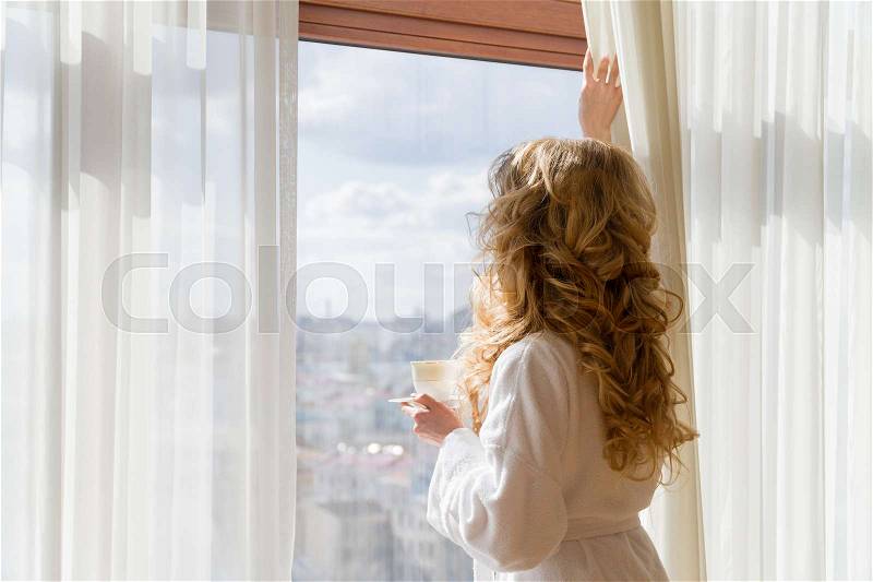 Beauty girl drinking coffee. Beautiful woman opening curtains, looking out the window and enjoying her morning coffee, stock photo