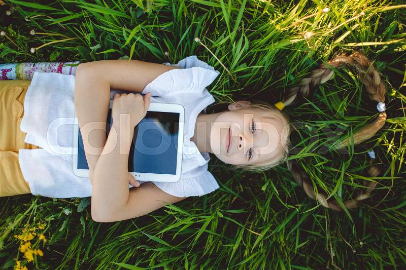 Little cute blonde girl is lying on back with digital tablet in yellow field with flowers, stock photo