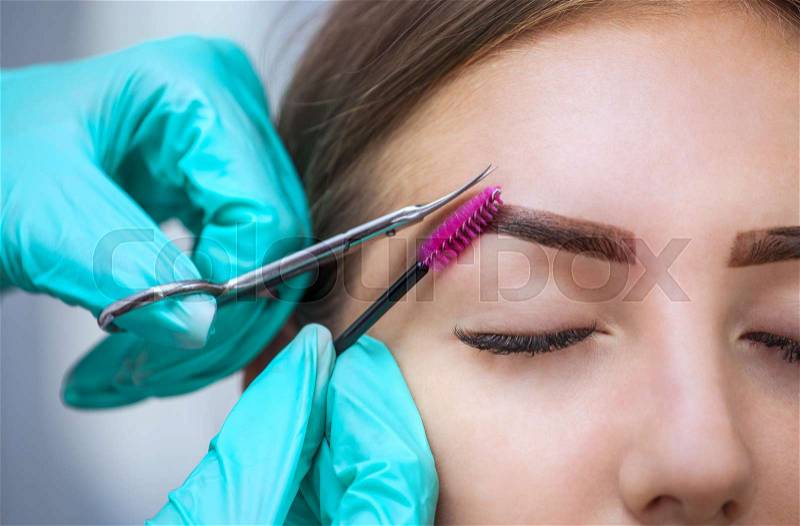 Master makeup corrects, gives shape to and cut with scissors previously painted with henna eyebrows in a beauty salon. Professional care for face, stock photo