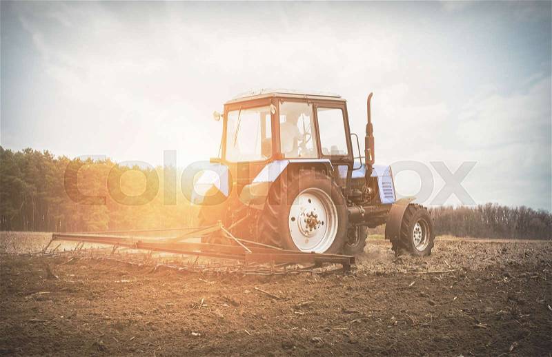 In the early spring morning because of the wood the bright sun ascends.The tractor goes and pulls a plowplowing a field before landing of crops, stock photo