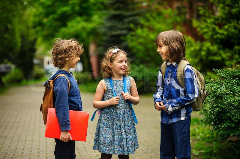 Cute little school students briskly talk on the schoolyard. Children have a good mood. Warm spring morning. Behind shoulders at schoolmates schoolbags. In hands of one of boys bright red folder, stock photo
