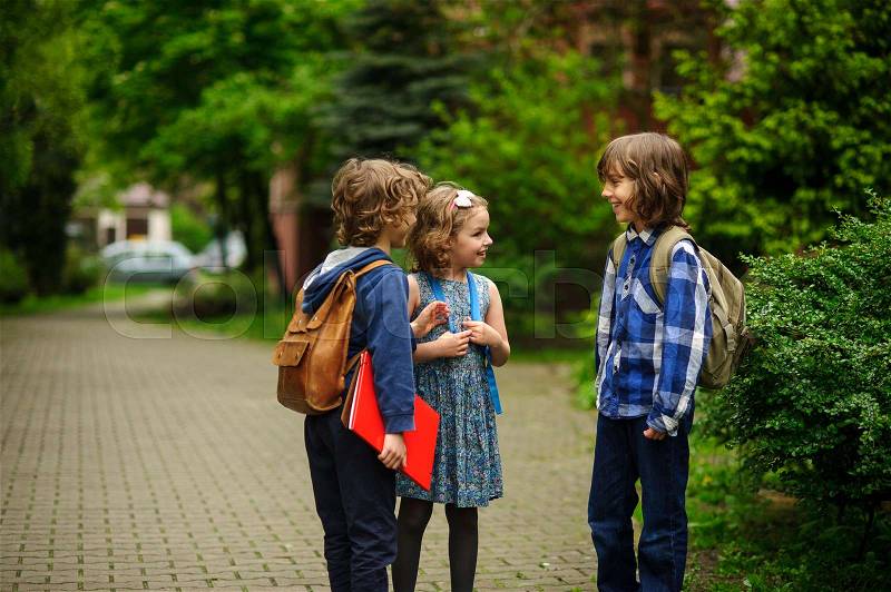 Little school students briskly talk on the schoolyard. Children have a good mood. Warm spring morning. Behind shoulders at schoolmates schoolbags, stock photo