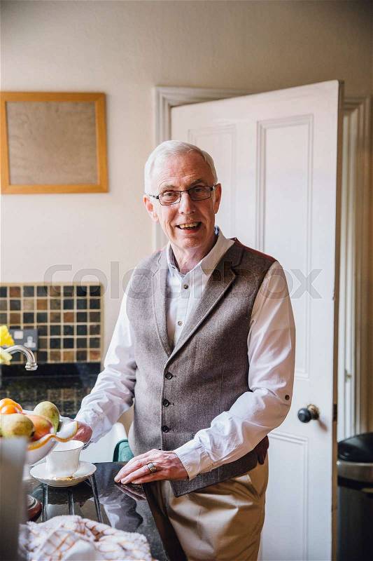 Happy senior man is smiling for the camera while standing in his kitchen with a cup of tea, stock photo