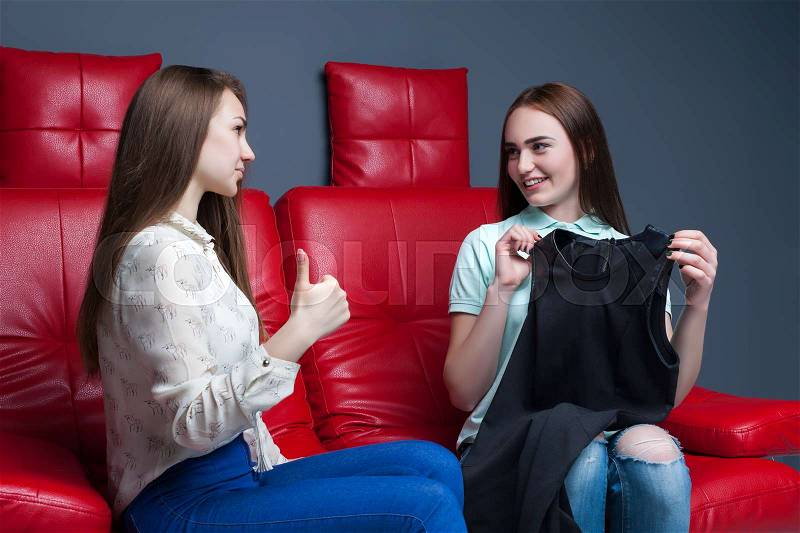 Two women sitting on red leather couch and tries on dresses. Leisure of happy girlfriends. Female friendship, stock photo