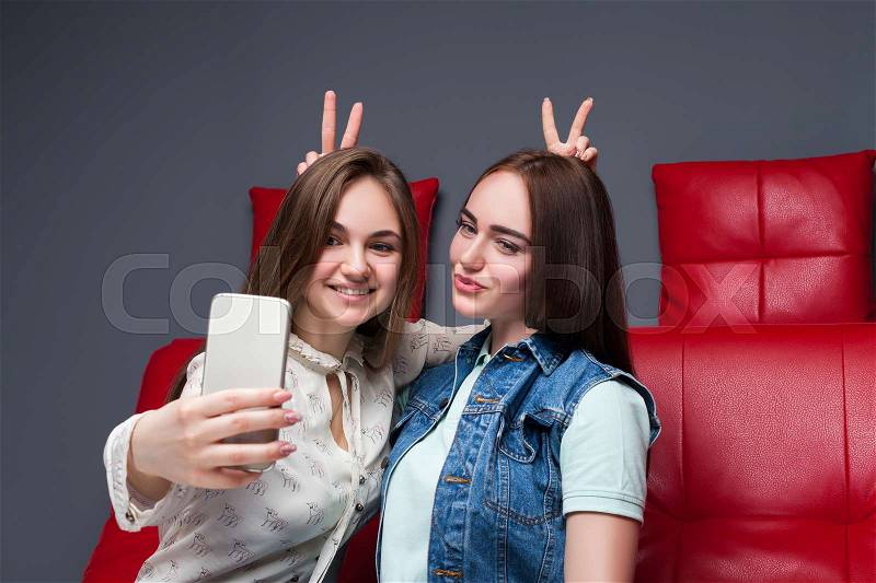 Two funny girlfriends sitting on red leather couch and makes selfie on phone camera. Female friendship. Leisure of happy girls, stock photo