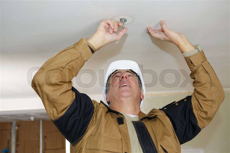 Middle-age electrician fixes halogen lamp in ceiling light in room, stock photo