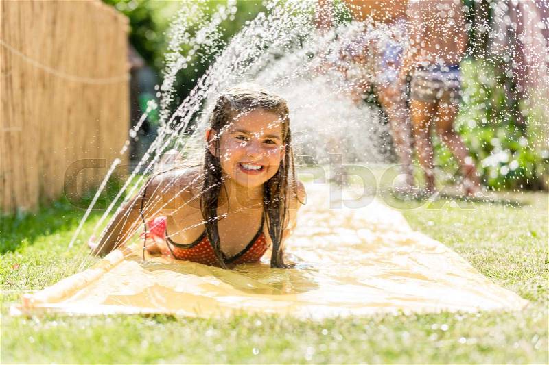 Water fun in garden - girl cooling down with water sprinkler on garden slide, stock photo