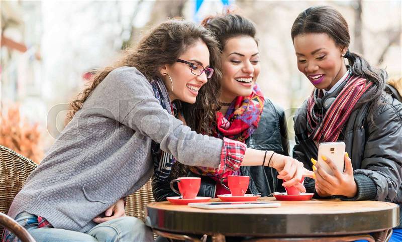Multicultural group of women in cafe showing each other pictures on smart phone and chatting, stock photo