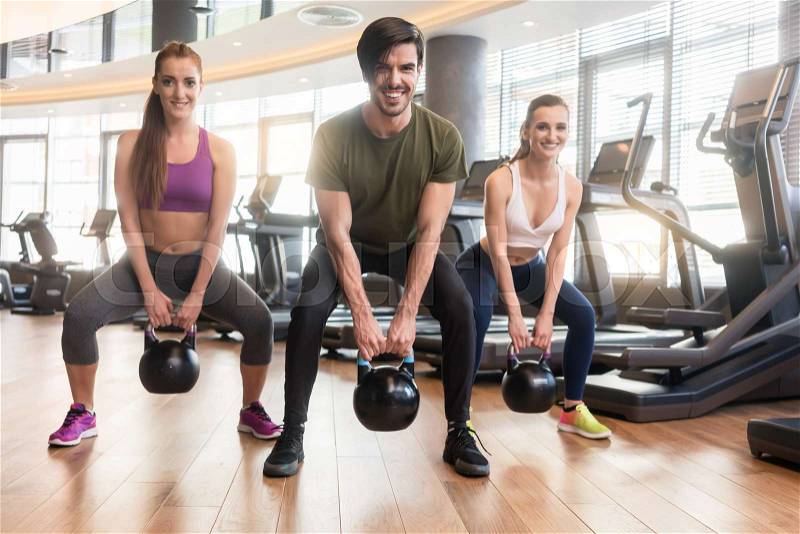 Three fit young people smiling and looking at camera while exercising kettlebell swings during full body workout at the gym, stock photo