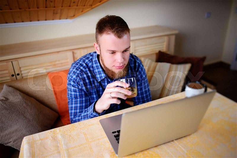 Young professional surfing the Internet on his laptop and drinking whiskey from a glass, stock photo