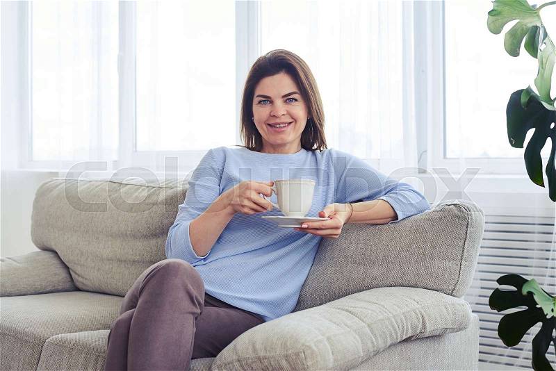 Mid shot of smiling madam having rest on sofa with cup of coffee, stock photo