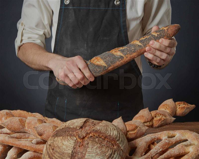Baker holds a baguette with poppy seeds on a dark background and a variety of bread on the table, stock photo