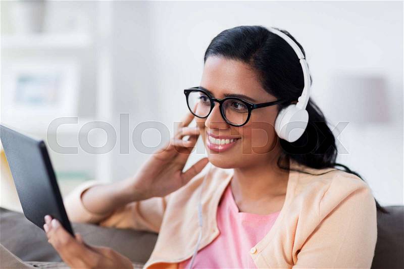 People, technology and leisure concept - happy young woman sitting on sofa with tablet pc computer and headphones listening to music at home, stock photo