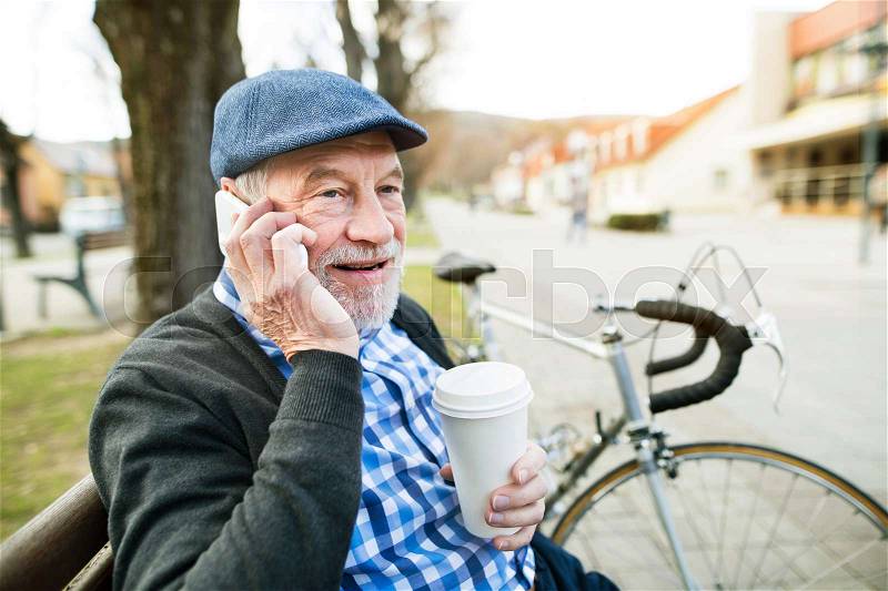 Handsome senior man with bicycle in town park sitting on bench, holding coffee and smart phone, making phone call. Sunny spring day, stock photo