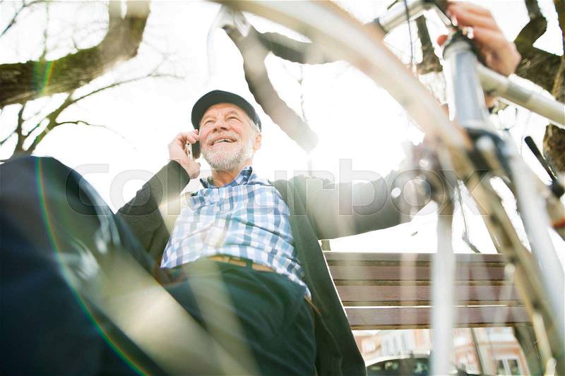 Handsome senior man with bicycle in town park sitting on bench with legs crossed, holding smart phone, making phone call. Sunny spring day, stock photo