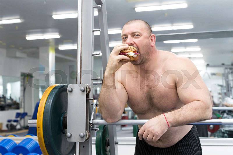 Big fat hungry man chewing a hamburger with meat and cheese in the gym, stock photo
