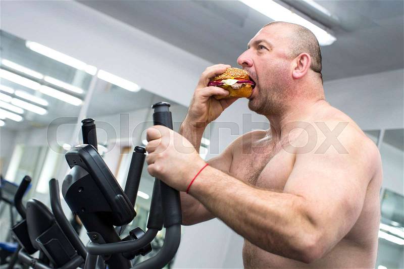 Big fat hungry man chewing a hamburger with meat and cheese in the gym, stock photo