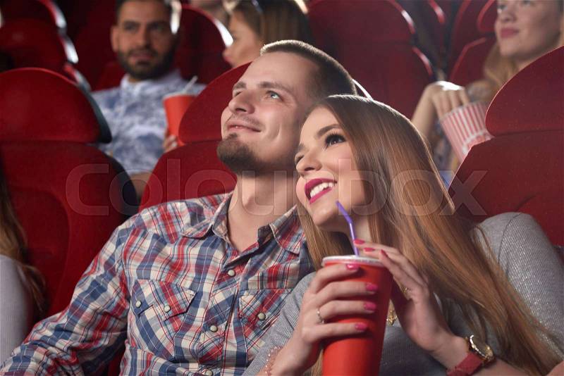 Smiling beautiful couple embracing, drinking cola, watching interesting movie at modern cinema. Boyfriend and girlfriend sitting in big cinema hall, girl leaning on her boy, looking at projector, stock photo