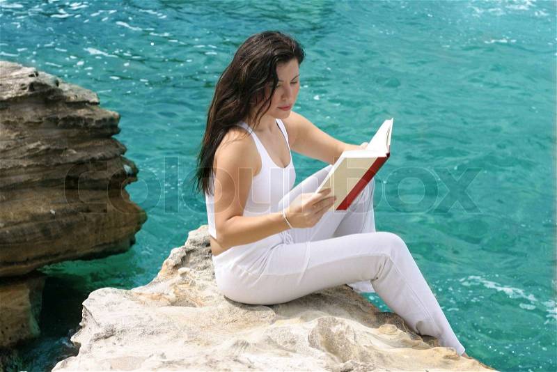 A beautiful female woman relaxes by ocean seaside reading a book. Focus on woman only, stock photo