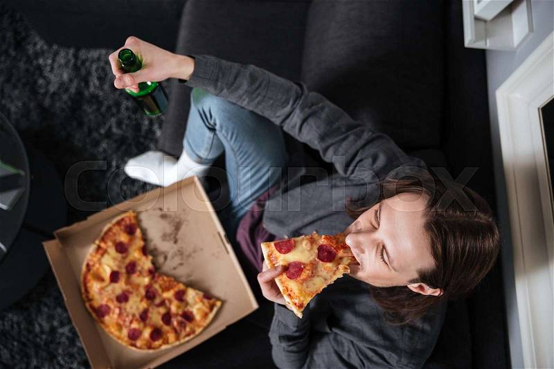 Image of young man sitting at home indoors eating pizza. Watch TV while looking aside, stock photo