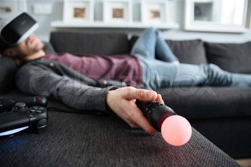 Image of young man lies on sofa at home indoors wearing 3d virtual reality glasses holding joystick. Focus on joystick, stock photo