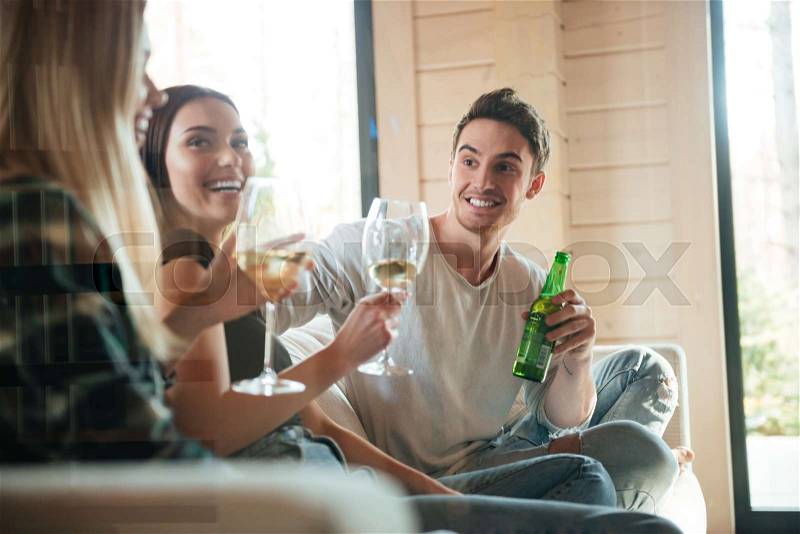 Group of cheerful young friends drinking wine and beer on sofa at home, stock photo