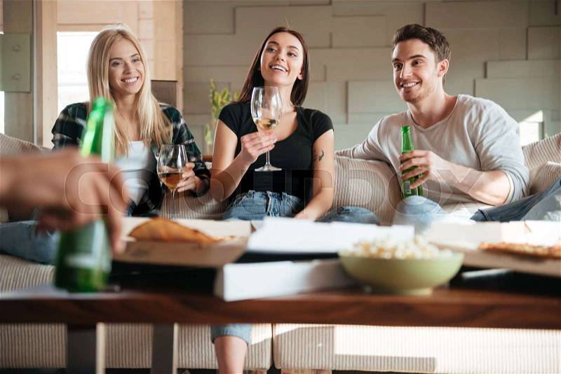 Group of smiling young friends with pizza, wine and beer sitting and talking on sofa, stock photo