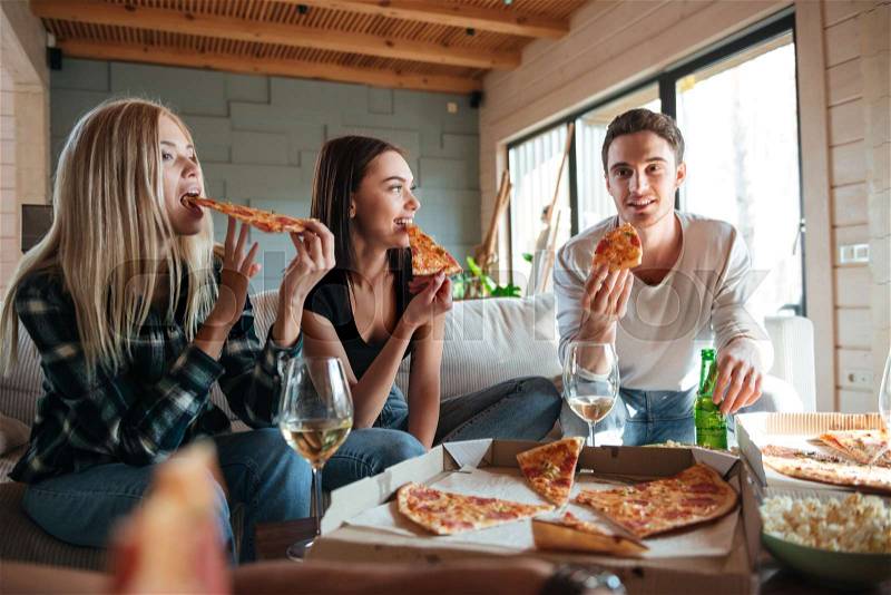 Three friends are sitting on sofa and eating pizza in house, stock photo