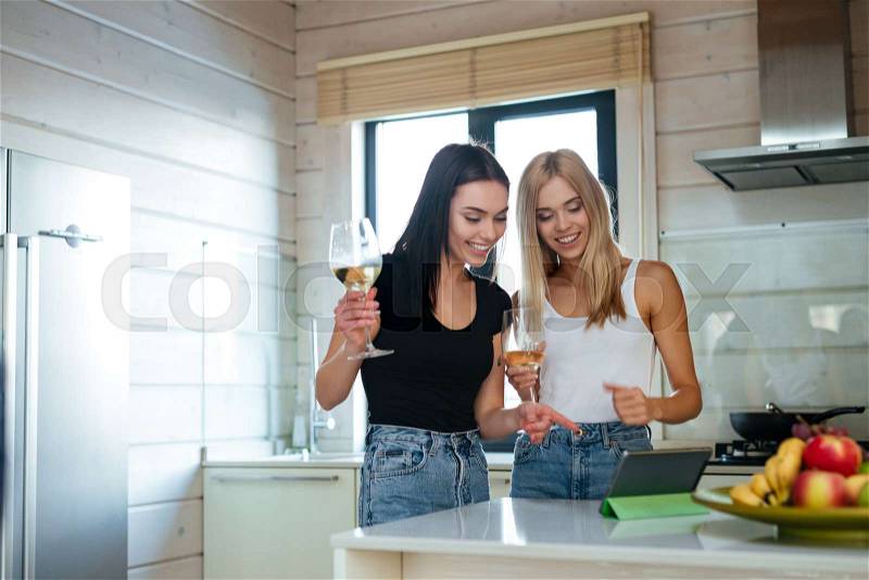 Two happy women drinking wine and using tablet computer on the kitchen, stock photo