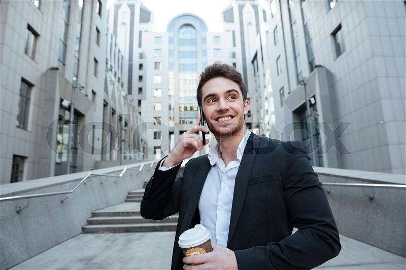Smiling businessman holding coffee and talking on phone while walking, stock photo