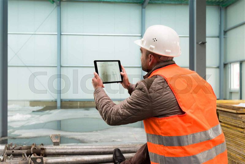Construction supervisor checks the interior of a new warehouse being constructed with a digital tablet showing a map in his hand, wearing a safety helmet and vest, stock photo