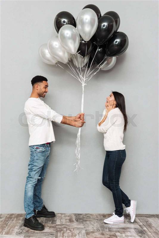 Image of young smiling loving couple standing over grey wall. Man give to woman a lot of balloons. Looking aside, stock photo